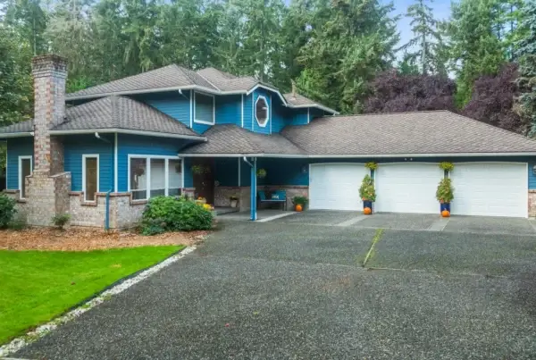 A two-story blue house with white trim, a large attached three-car garage, brick chimney, well-kept lawn, and trees in the background. Hanging plants and flower pots decorate the entrance and driveway.