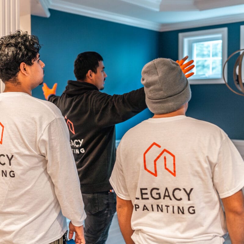 Three people wearing Legacy Painting shirts stand in a room with blue walls and white trim, facing windows. One person gestures toward the ceiling light fixture, discussing or inspecting the space.