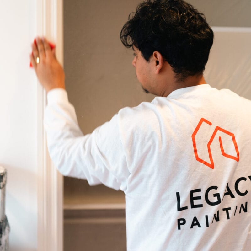 A person wearing a white “Legacy Painting” shirt sands a doorframe. Shelving and a step ladder are visible in the background.