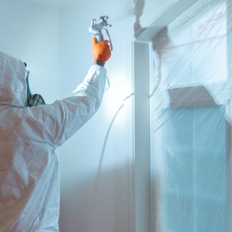 A person in protective gear and an orange glove uses a spray gun to paint a white wall near a window covered with plastic sheeting.