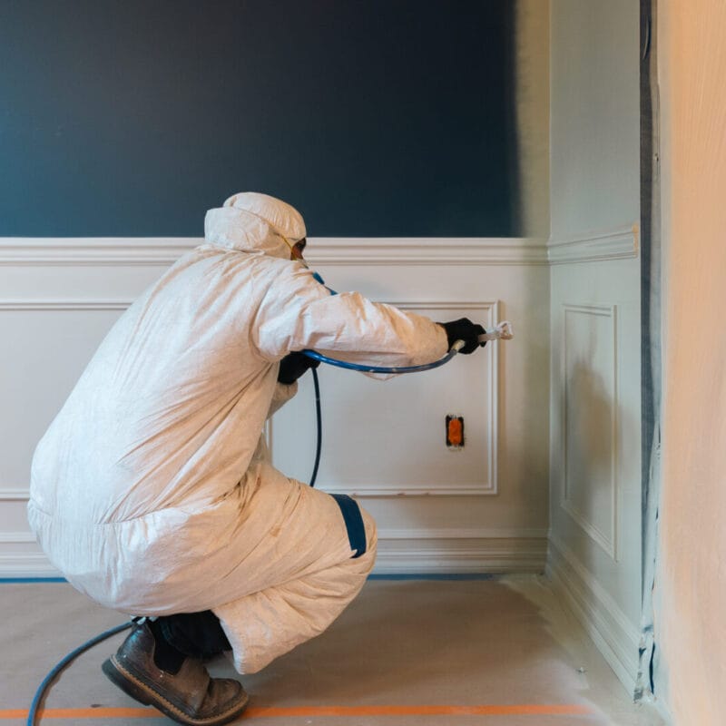 A person in protective coveralls uses a paint sprayer to paint a white paneled wall. The floor and nearby surfaces are covered for protection. The adjacent wall is dark blue.