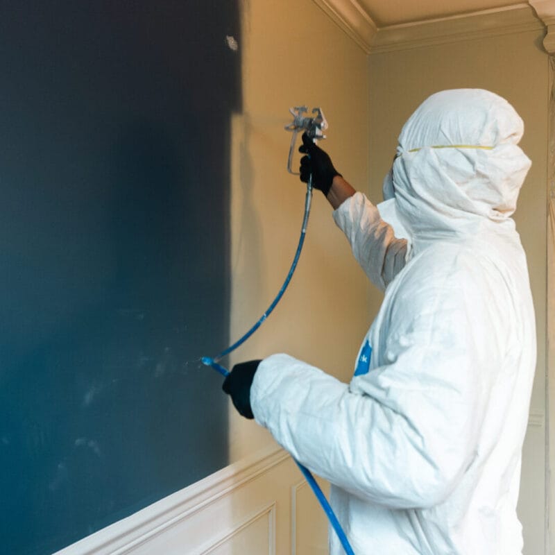 A person wearing protective clothing and gloves uses a spray gun to paint a wall indoors. One side of the wall is blue, while the other is yellow. Plastic sheeting covers part of the room for protection.