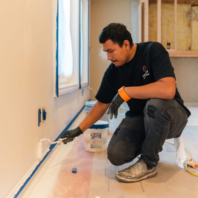 A man kneels on the floor and paints a wall with a roller in a room under renovation. He is wearing work clothes and gloves, with painting supplies and construction materials nearby.