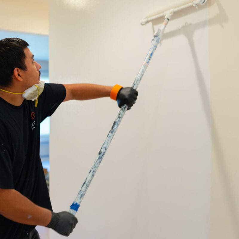 A person wearing gloves and a mask uses a roller brush to paint a white wall in a brightly lit room.