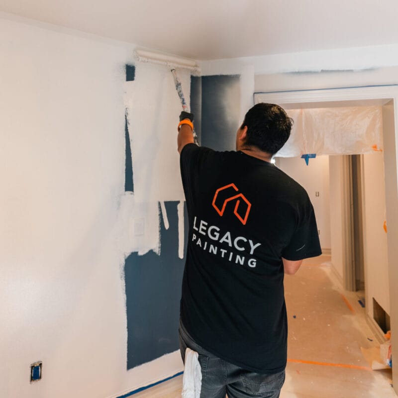 A person wearing a Legacy Painting shirt uses a roller to paint a white wall over dark blue paint in a room under renovation. Plastic covers the floor and door frame for protection.