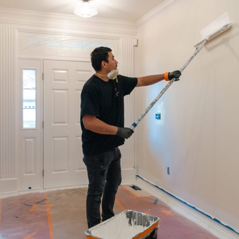 A person wearing gloves and a mask uses a paint roller to paint a white wall in a home interior. Floors and doorways are covered for protection, and painting supplies are visible nearby.