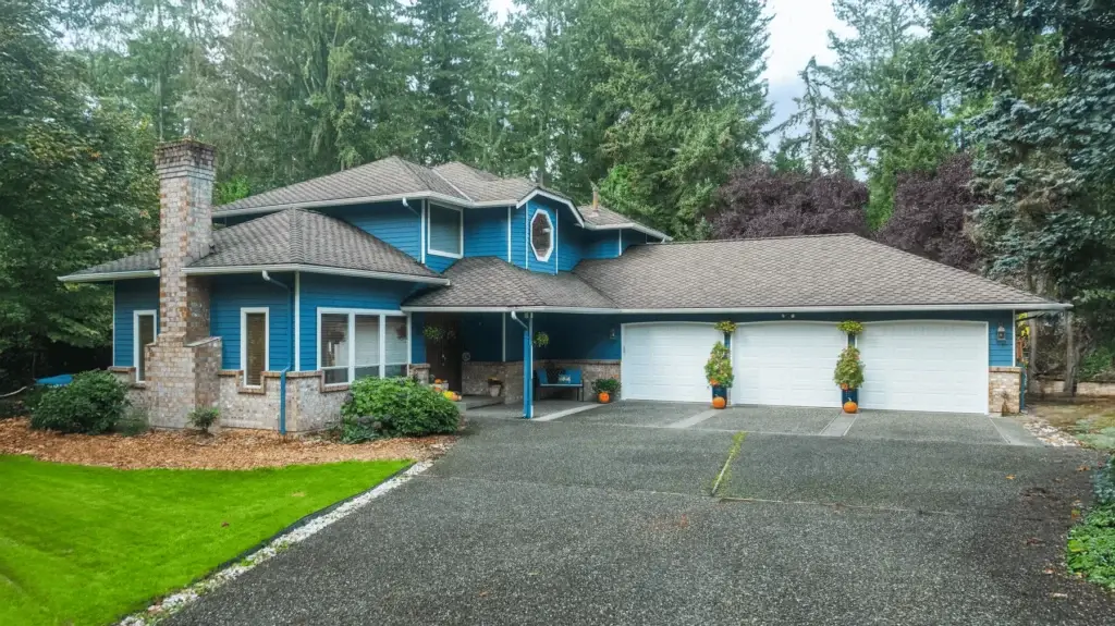 A two-story blue house with a large three-car garage, brick chimney, manicured lawn, and surrounded by tall trees and greenery. Four orange potted plants sit in front of the garage doors.