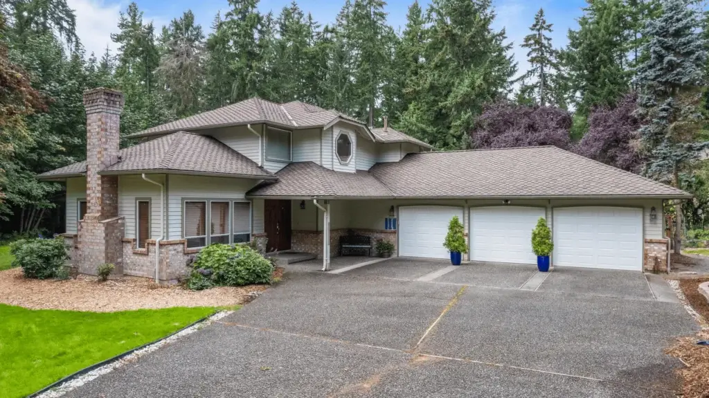 Two-story suburban house with a large driveway, three-car garage, and brick accents. Surrounded by lush greenery, trees, and neatly landscaped shrubs. The exterior is light with a shingled roof.